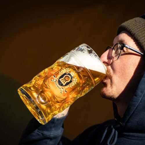 A person in glasses, a beanie, and a dark hoodie drinks beer from an MT. Nelson Glass with a crest logo, set against a dark background.