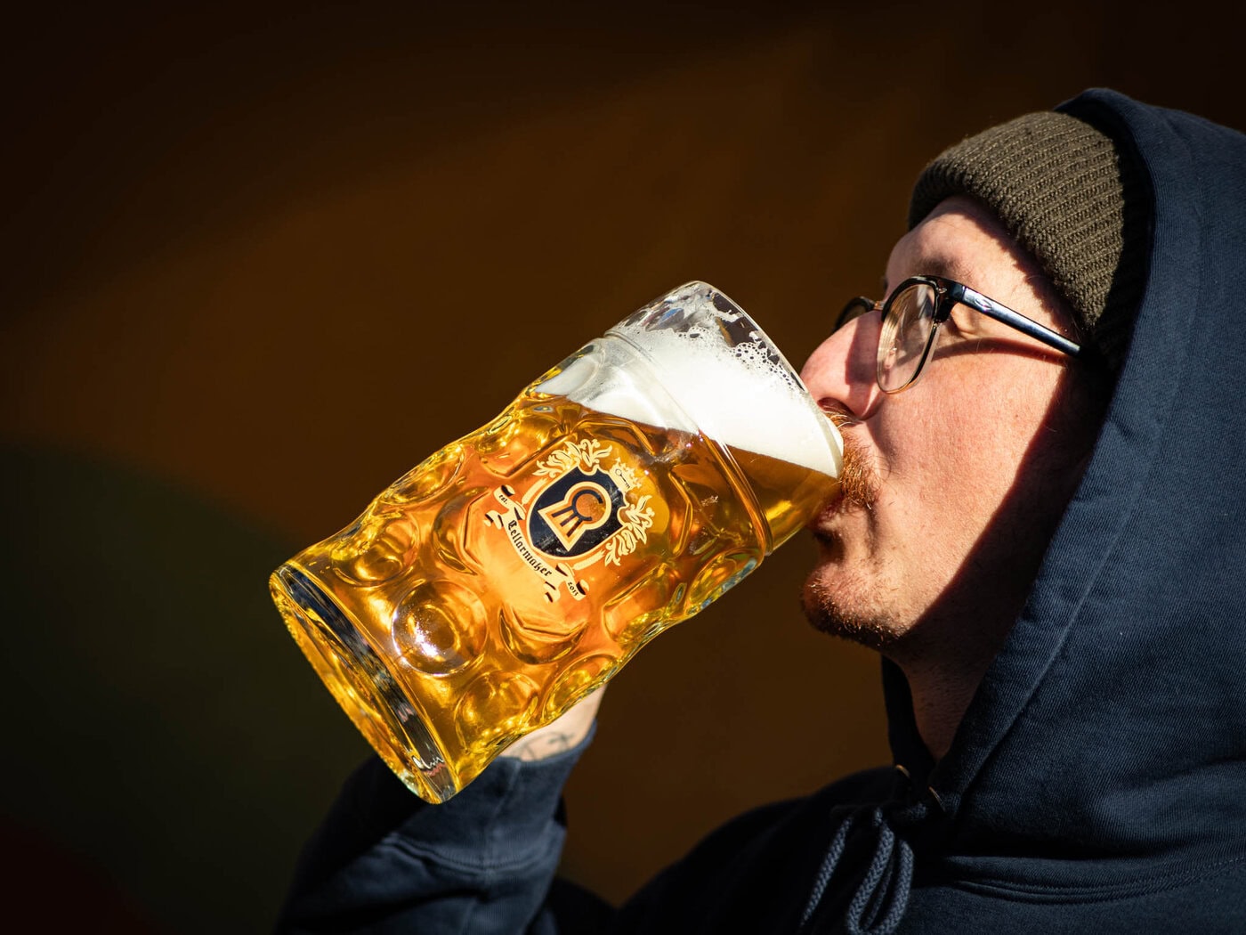 A person in glasses, a beanie, and a dark hoodie drinks beer from an MT. Nelson Glass with a crest logo, set against a dark background.