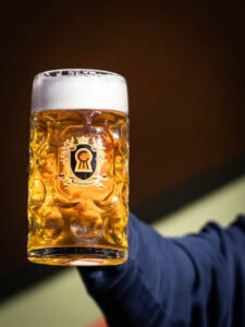 A person in a dark sleeve holds up the MT. Nelson Glass, a large clear mug filled with golden beer, thick white foam on top, and a decorative logo on the front.