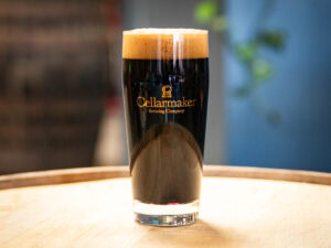 A MT. Nelson Glass filled with dark beer and thick, creamy foam sits on a wooden surface, branded with the Cellarmaker Brewing Company logo. The softly blurred background highlights the elegant presentation.