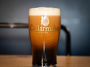 A MT. Nelson Glass filled with dark beer and creamy foam sits on a wooden surface, labeled "Cellarmaker Brewing Co." with a softly blurred background.