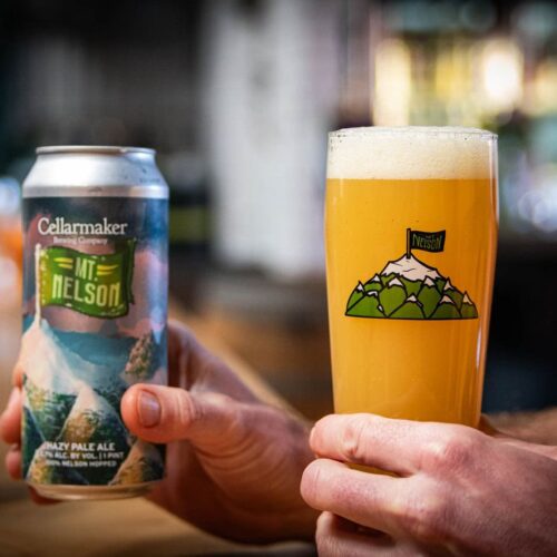 A person holds a can of pale ale in one hand and a pint of hazy, golden beer with a frothy head in the other, poured into the MT. Nelson Glass, which features a mountain illustration.