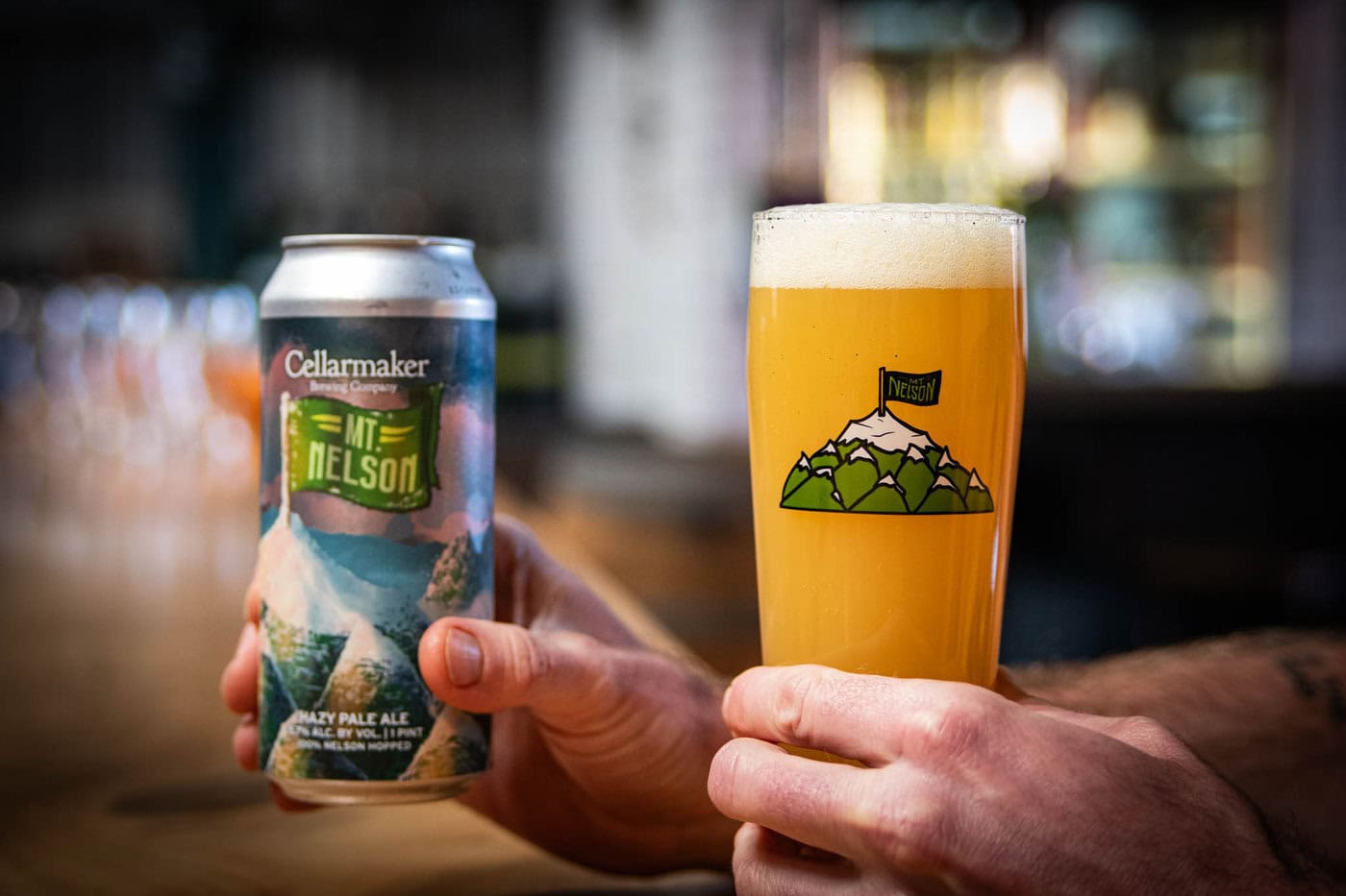 A person holds a can of pale ale in one hand and a pint of hazy, golden beer with a frothy head in the other, poured into the MT. Nelson Glass, which features a mountain illustration.