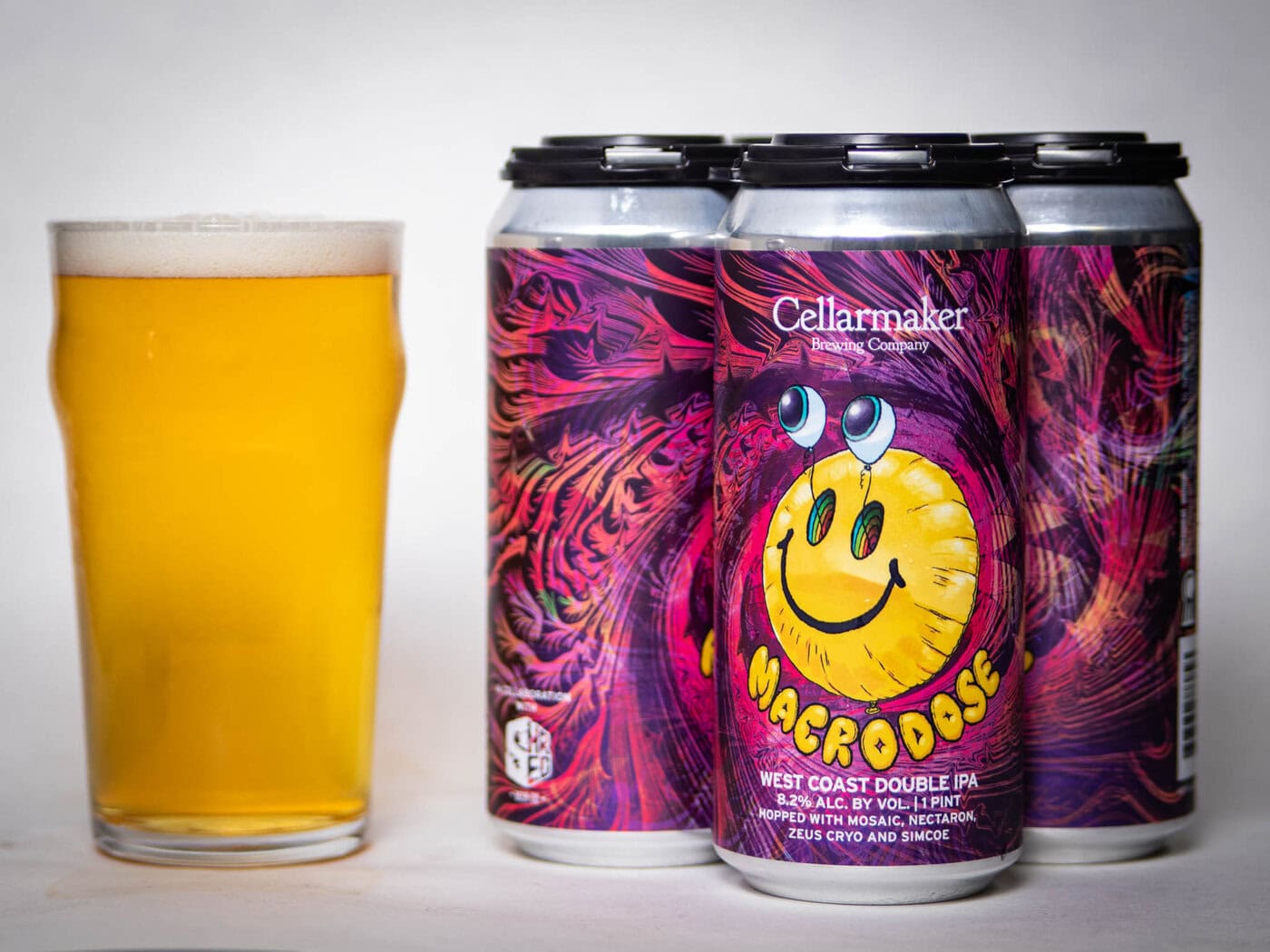 A Pint Glass Of Hazy Golden Beer Stands Next To Four Cans Of Cellarmaker’s “Macrodose” West Coast Double Ipa, Featuring A Vibrant, Psychedelic Label With A Yellow Smiley Face.