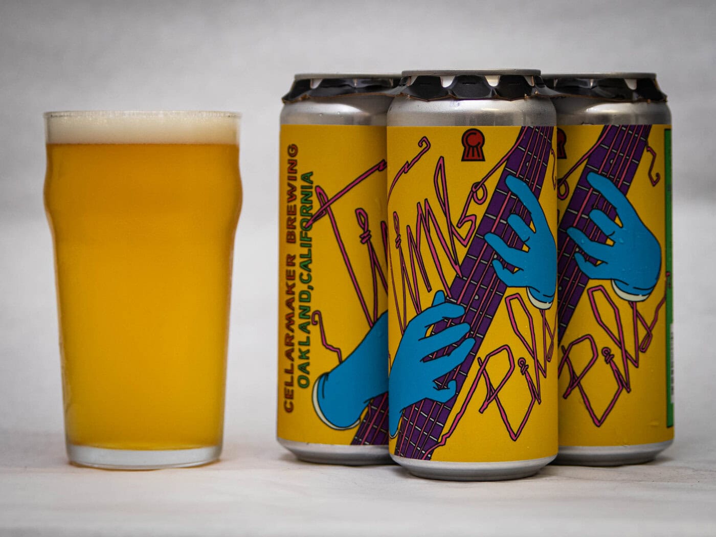 A Pint Glass Of Hazy, Golden Beer Stands Next To Three Cans With Yellow Labels Featuring Blue Hands Playing A Purple Guitar. The Cans Are Labeled &Quot;Cellarmaker Brewing, Oakland, California.