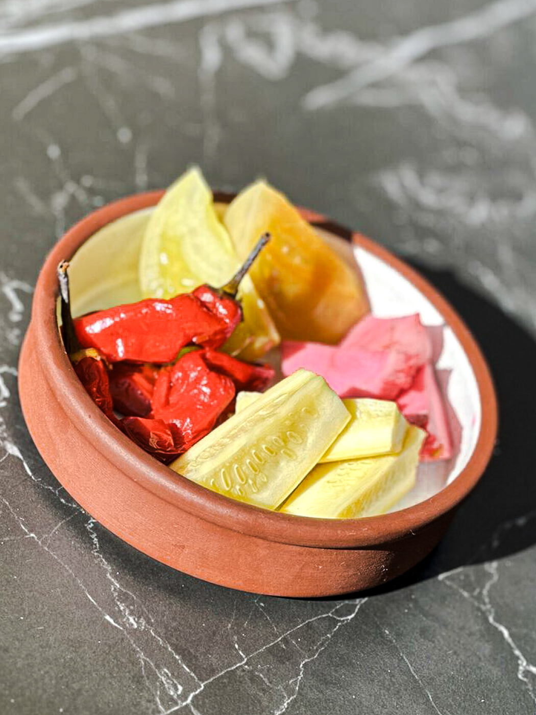 A Small Terracotta Bowl Holds Assorted Pickled Vegetables, Including Sliced Cucumbers, Bright Red Peppers, Yellow Pickles, And Pink Pickled Ginger, On A Dark Marble Surface.