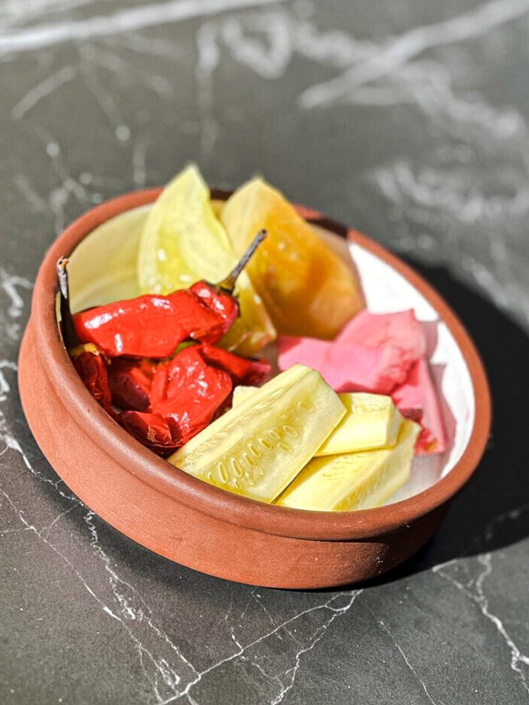 A Small Terracotta Bowl Holds Assorted Pickled Vegetables, Including Sliced Cucumbers, Bright Red Peppers, Yellow Pickles, And Pink Pickled Ginger, On A Dark Marble Surface.
