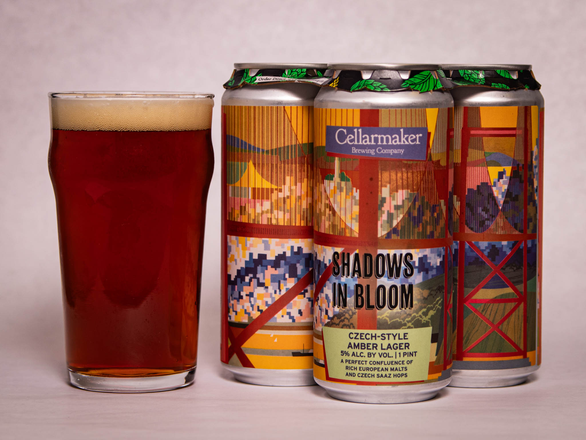 A Pint Glass Of Amber Beer Sits Next To Three Cans Of Cellarmaker Brewing Company'S &Amp;Quot;Shadows In Bloom&Amp;Quot; Czech-Style Amber Lager, Featuring Colorful, Abstract Artwork On The Cans.