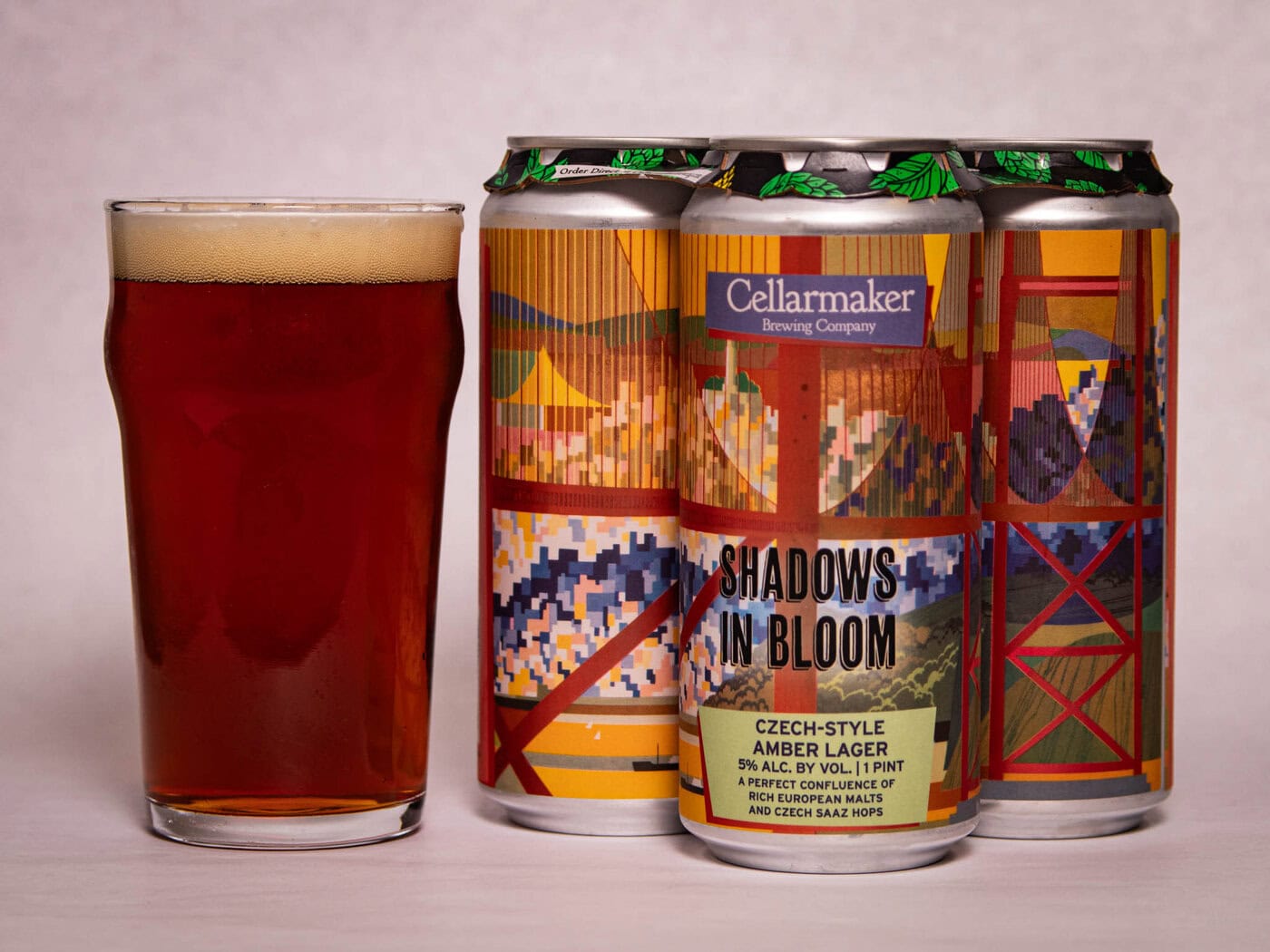 A Pint Glass Of Amber Beer Sits Next To Three Cans Of Cellarmaker Brewing Company'S &Quot;Shadows In Bloom&Quot; Czech-Style Amber Lager, Featuring Colorful, Abstract Artwork On The Cans.