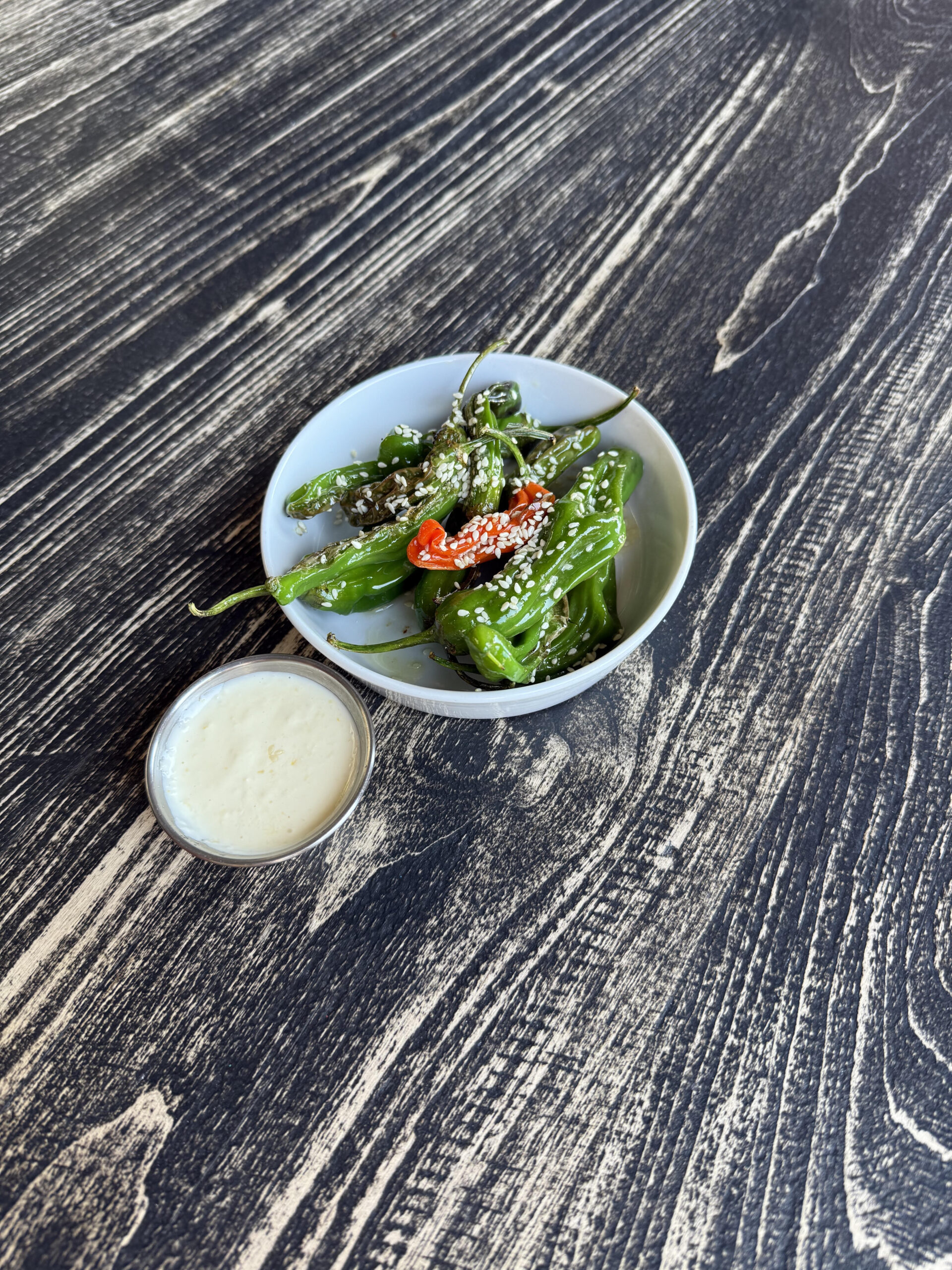 A small white bowl of blistered shishito peppers garnished with sesame seeds and sliced red chili, next to a small cup of creamy dipping sauce, on a dark wooden table.