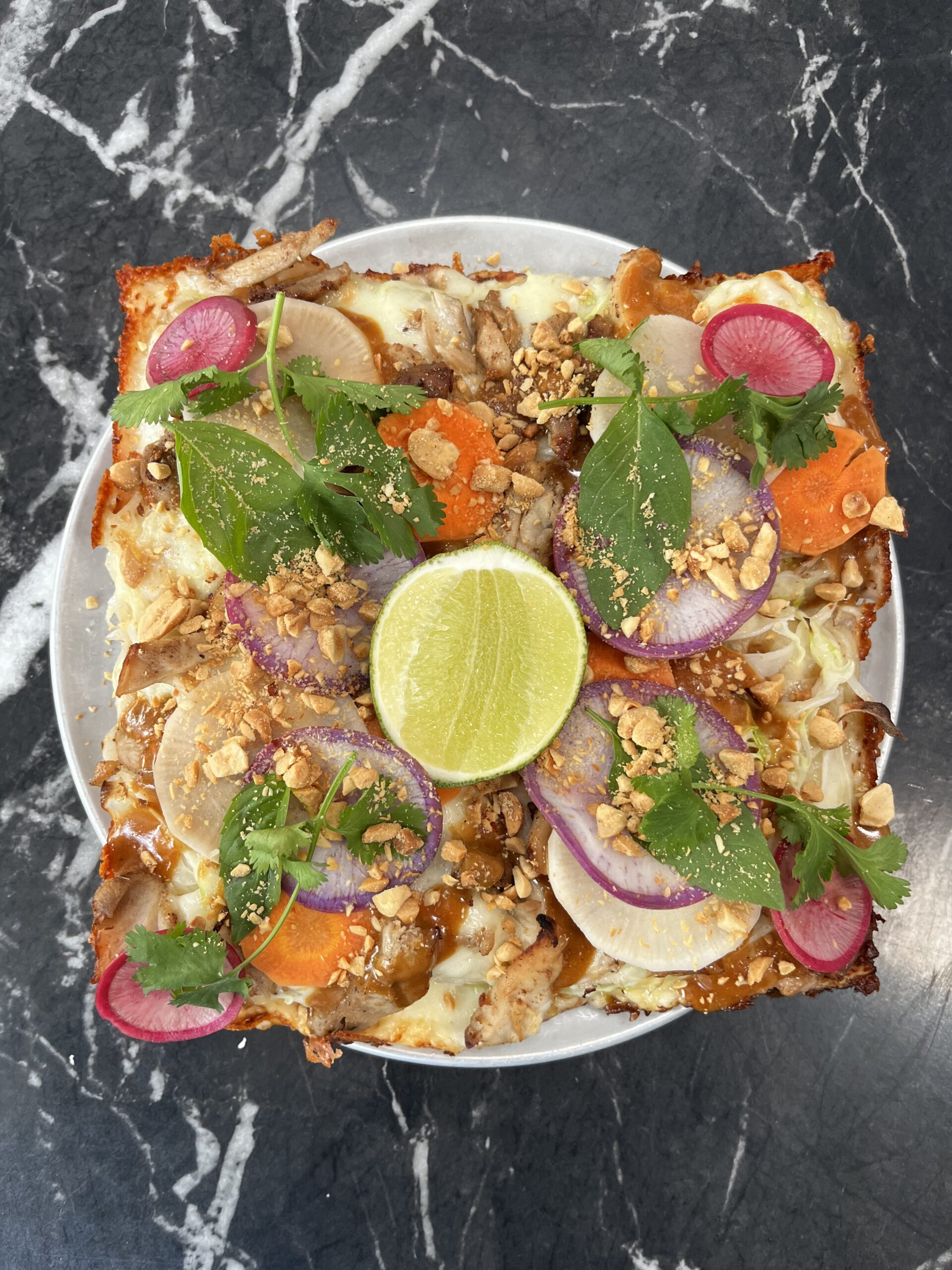 A colorful dish on a round white plate, topped with sliced radishes, red onions, carrots, herbs, crushed peanuts, and a halved lime, all on a dark marble surface.