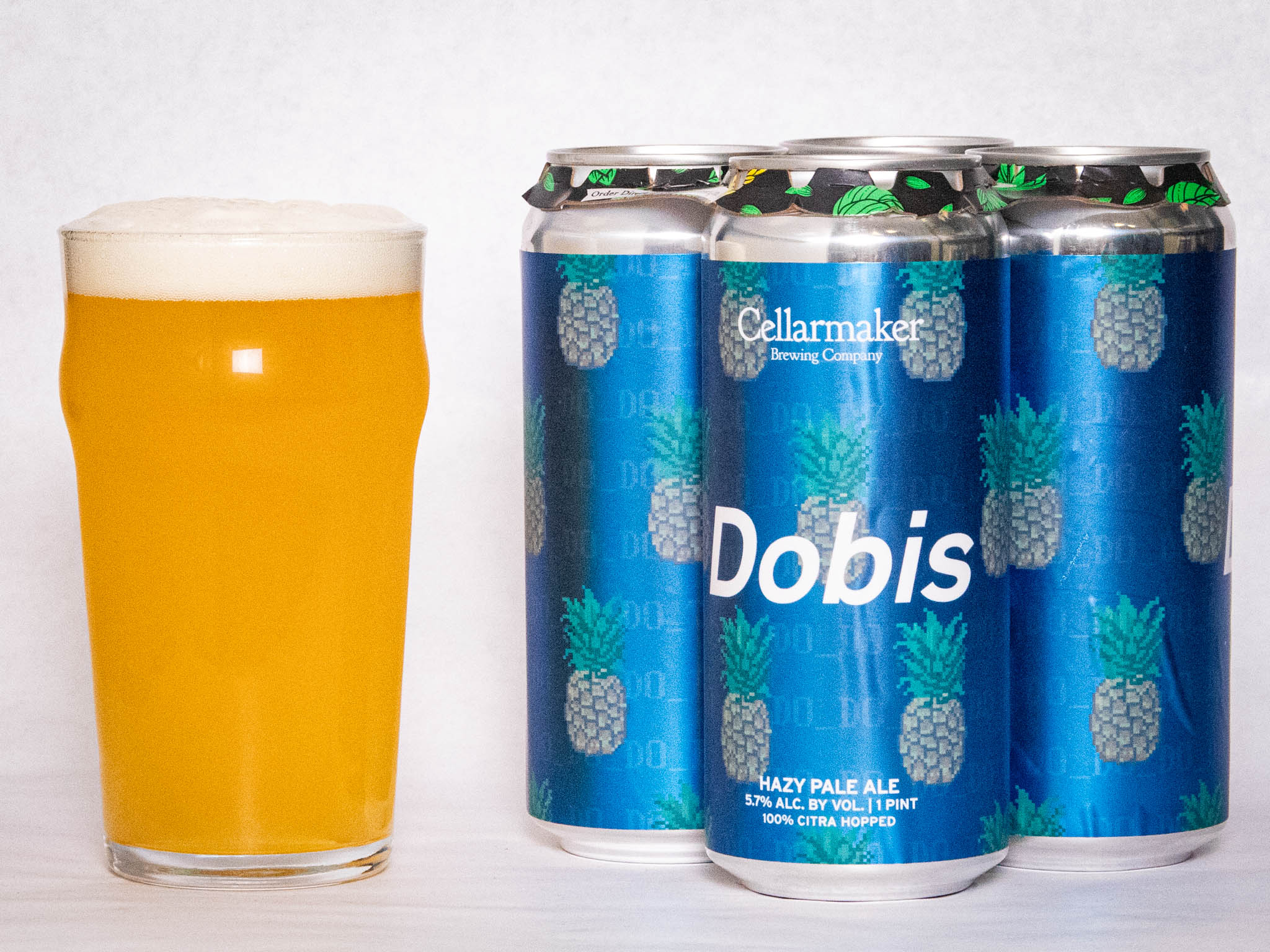 A Pint Glass Of Hazy Pale Ale Sits Next To A Four-Pack Of Cellarmaker Brewing Co. Dobis Cans, Which Are Blue With A Repeating Pineapple Design And White Text.