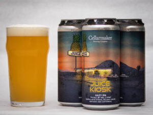 A pint glass filled with hazy golden beer sits next to four cans of Cellarmaker’s Juice Kiosk Hazy IPA, which feature artwork of a retro roadside kiosk with pineapples and desert mountains in the background.