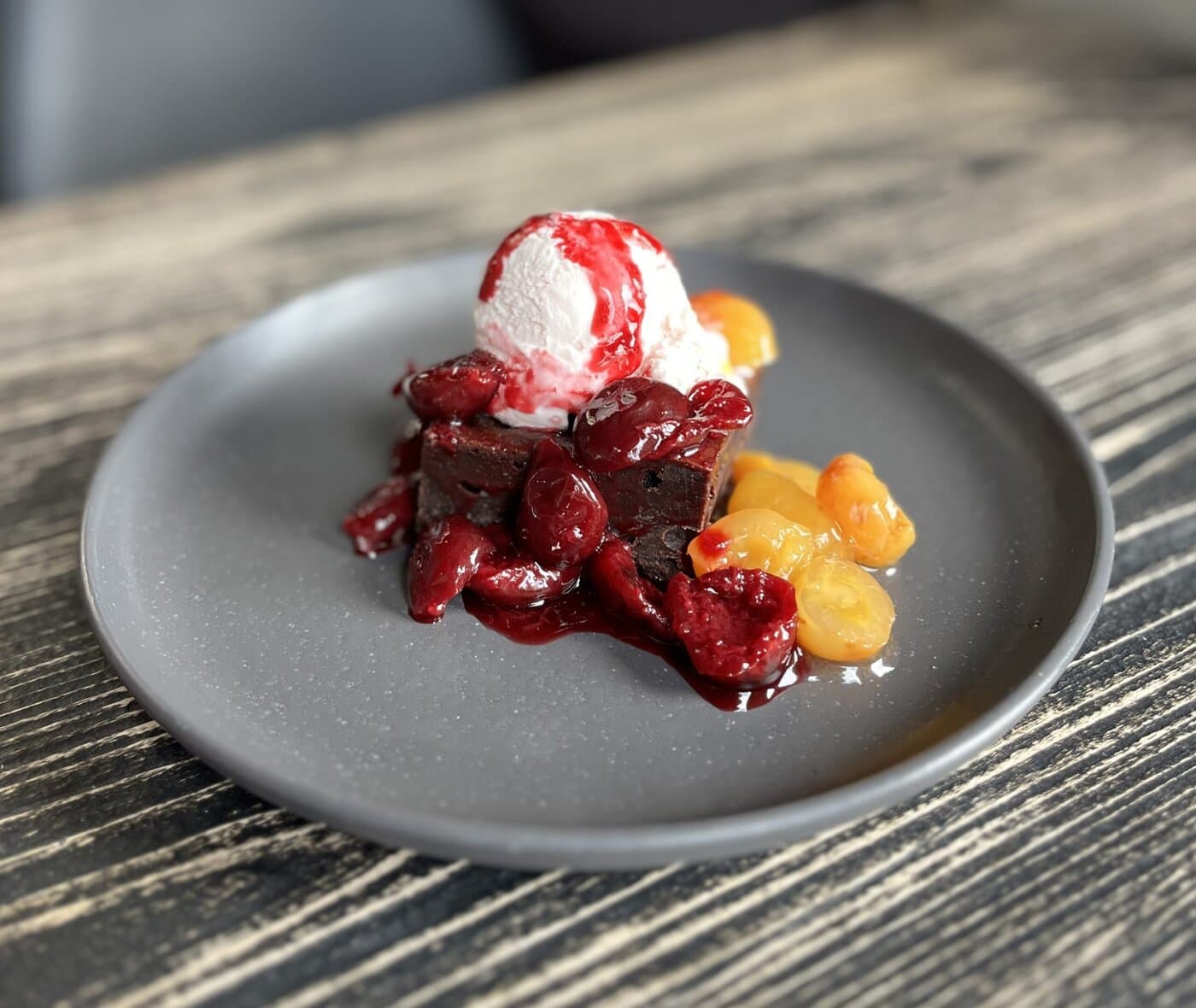 A chocolate brownie topped with vanilla ice cream and red berry sauce, served with red and yellow cherry compote, on a dark gray plate placed on a wooden table.