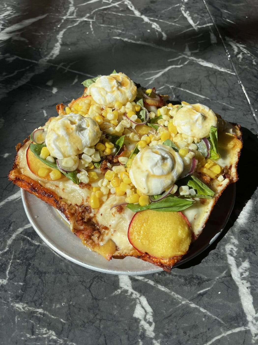 A square slice of pizza topped with corn, sliced peaches, onions, green vegetables, and four dollops of creamy white cheese, served on a small round plate on a dark marble surface.