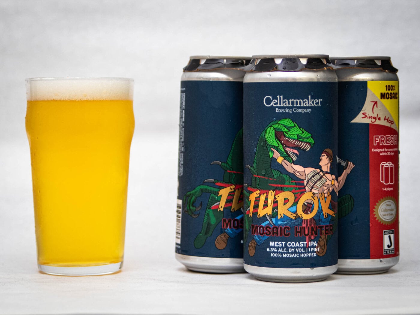 A Glass Of Hazy Yellow Beer Sits Next To Three Cans Of Cellarmaker Brewing Company’s “Turok Mosaic Hunter” West Coast Ipa, Featuring Colorful Artwork Of A Warrior Fighting A Dinosaur.