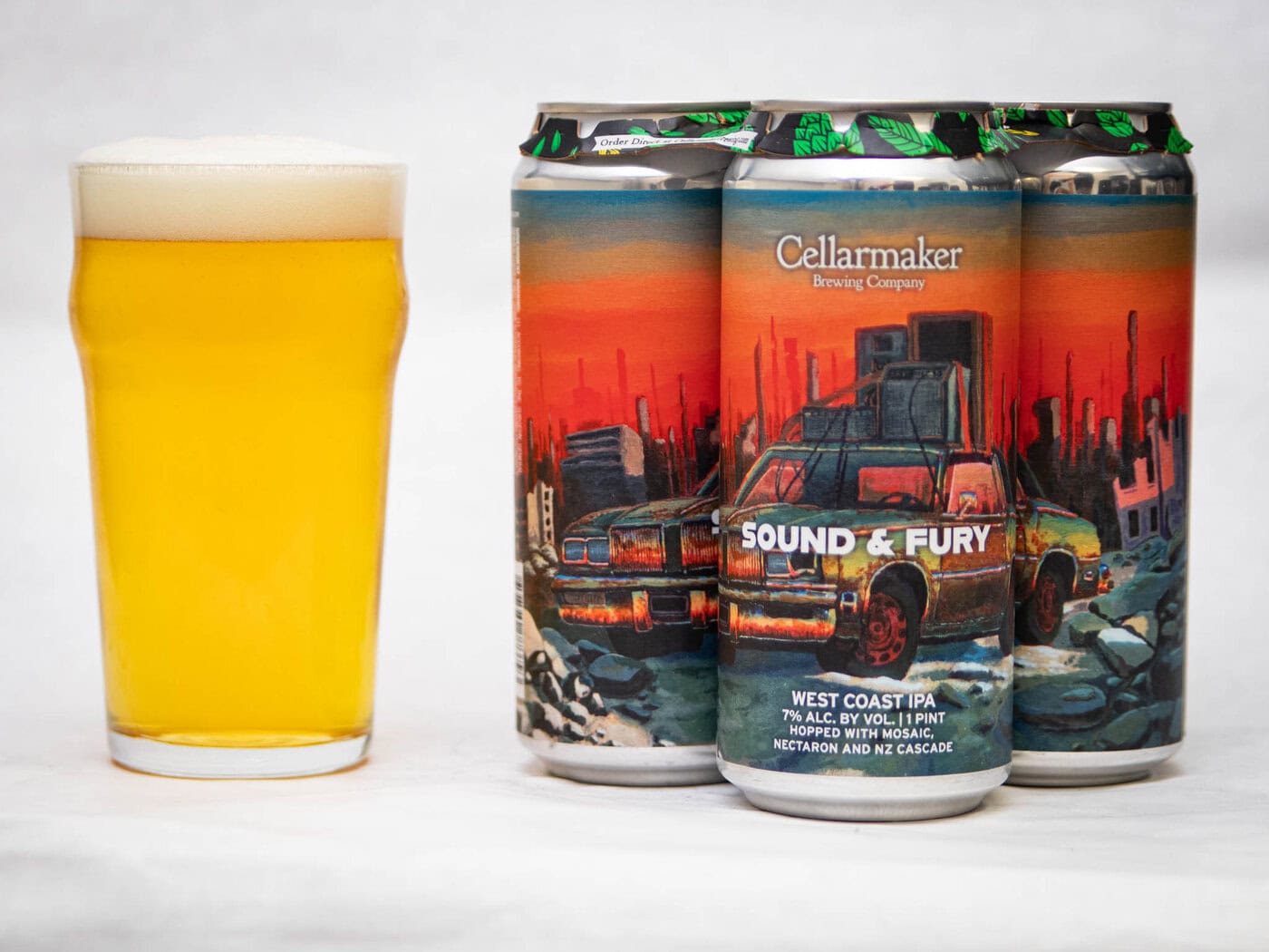 A Pint Glass Of Hazy Yellow Beer Sits Next To Three Cans Of Cellarmaker Brewing Company'S &Quot;Slow Train&Quot; West Coast Pale Ale, Showcasing Colorful Artwork Of A Ruined City And An Old Car.