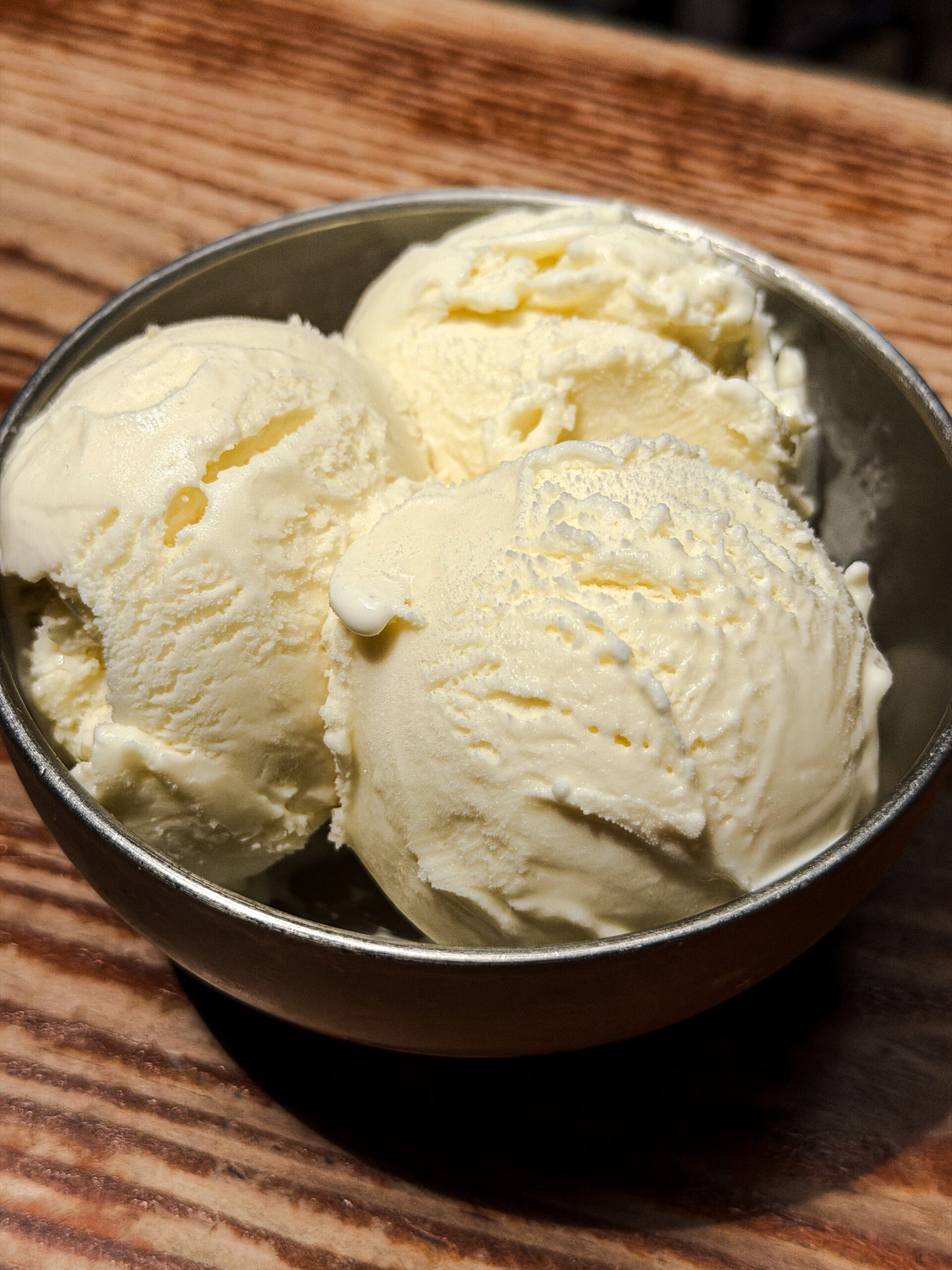 Three scoops of vanilla ice cream in a metal bowl, placed on a wooden surface.