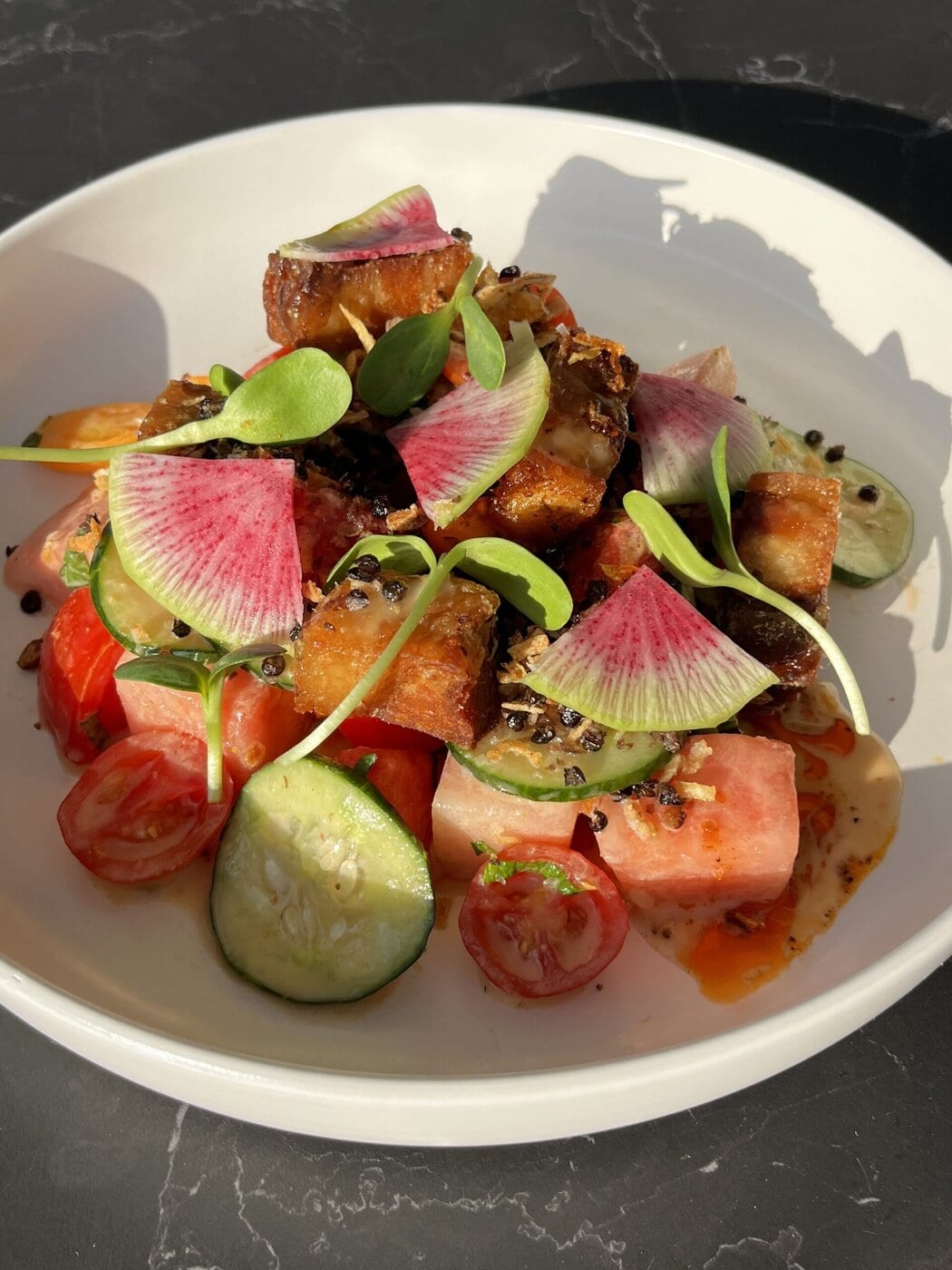 A white bowl with a colorful salad of sliced cucumbers, cherry tomatoes, watermelon cubes, seared pork belly, sliced radishes, microgreens, and a creamy dressing, all set on a dark marble surface.