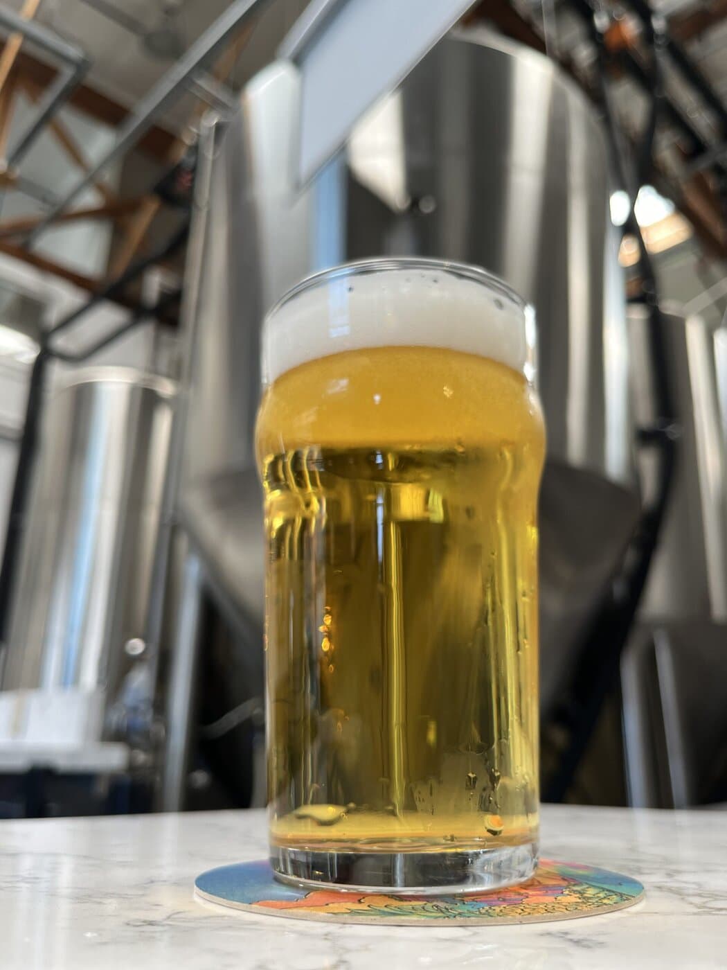 A Glass Of Pale Beer With A Foamy Head Sits On A Colorful Coaster Atop A White Table, With Large Stainless Steel Brewing Tanks Blurred In The Background.