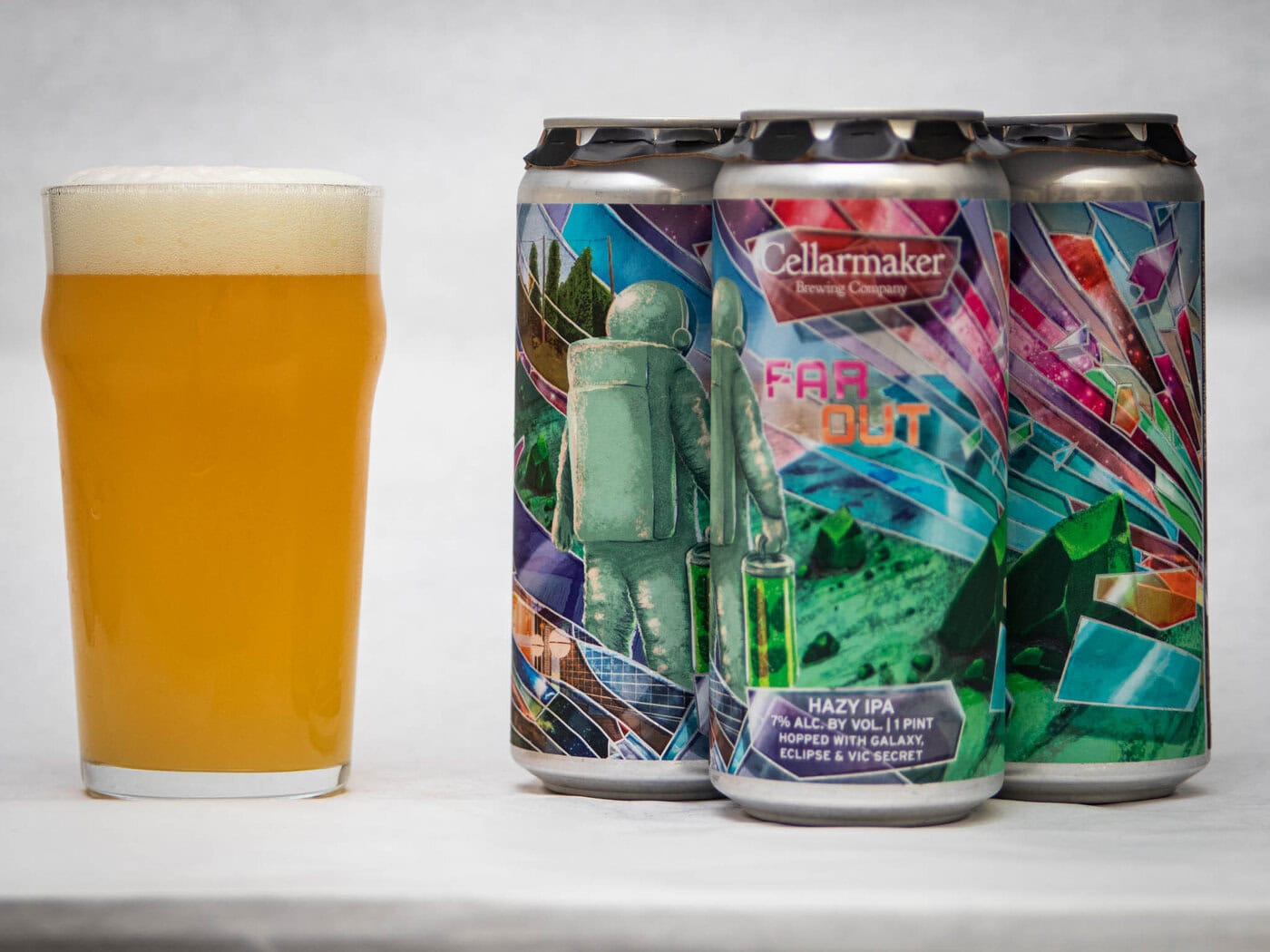 A Pint Glass Of Hazy Beer Stands Next To Three Colorful Cans Of Cellarmaker Brewing Company’s “Far Out” Hazy Ipa, Featuring Vibrant, Abstract Artwork With An Astronaut And Geometric Shapes.
