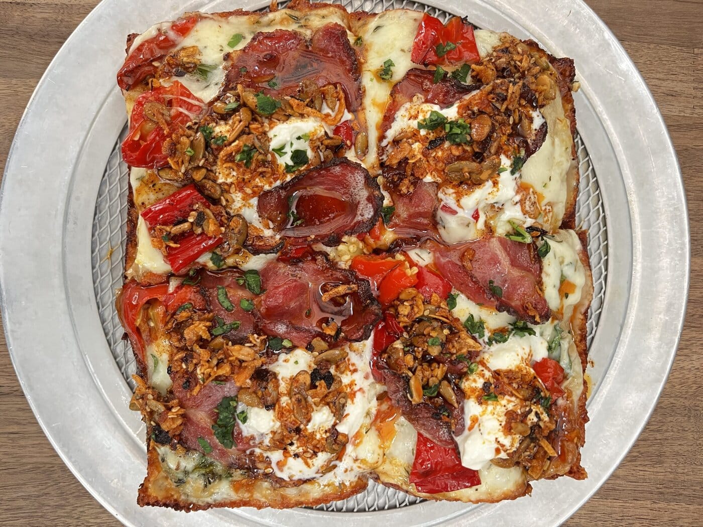 A rectangular pizza cut into six pieces, topped with melted cheese, sliced cured meat, chopped tomatoes, herbs, and a crunchy mixture, served on a round silver tray placed on a wooden table.