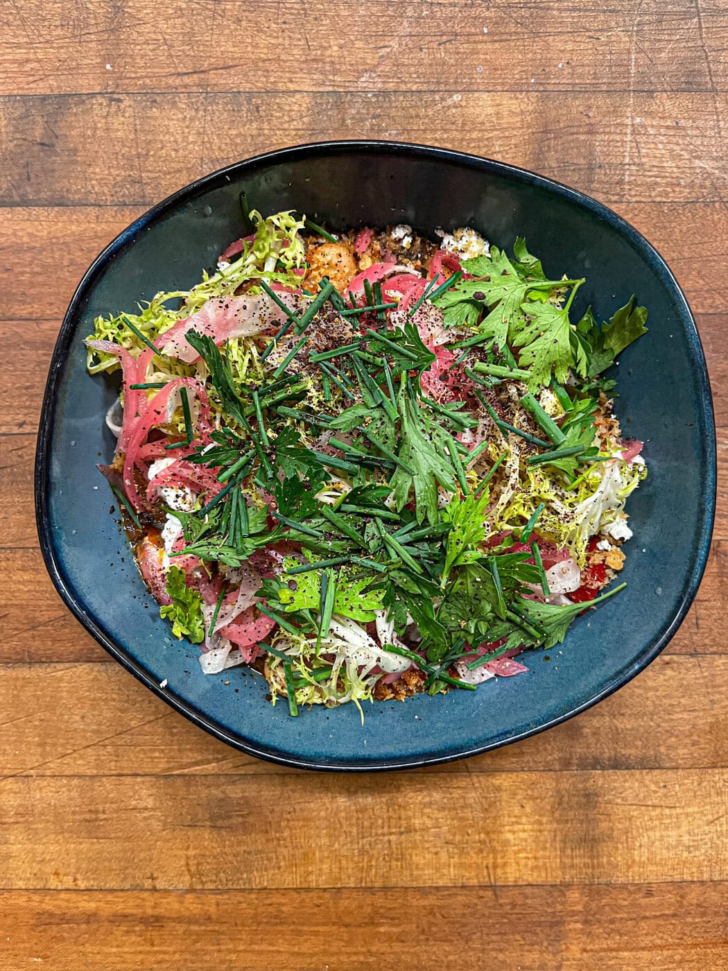 A Bowl Filled With A Colorful Salad Topped With Fresh Chopped Herbs, Pickled Onions, And Mixed Greens, Placed On A Wooden Table.