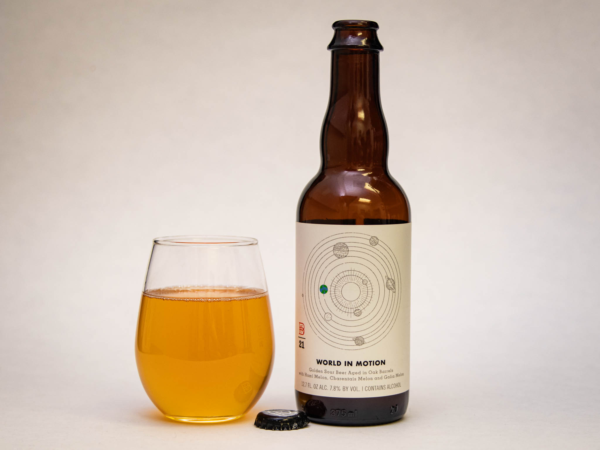 A Glass Of Golden Amber Beverage Sits Next To An Amber Bottle Labeled &Amp;Quot;World In Motion,&Amp;Quot; Featuring A Planetary Orbital Design On The Label. The Bottle Cap Rests In Front Of The Glass And Bottle On A Plain Background.
