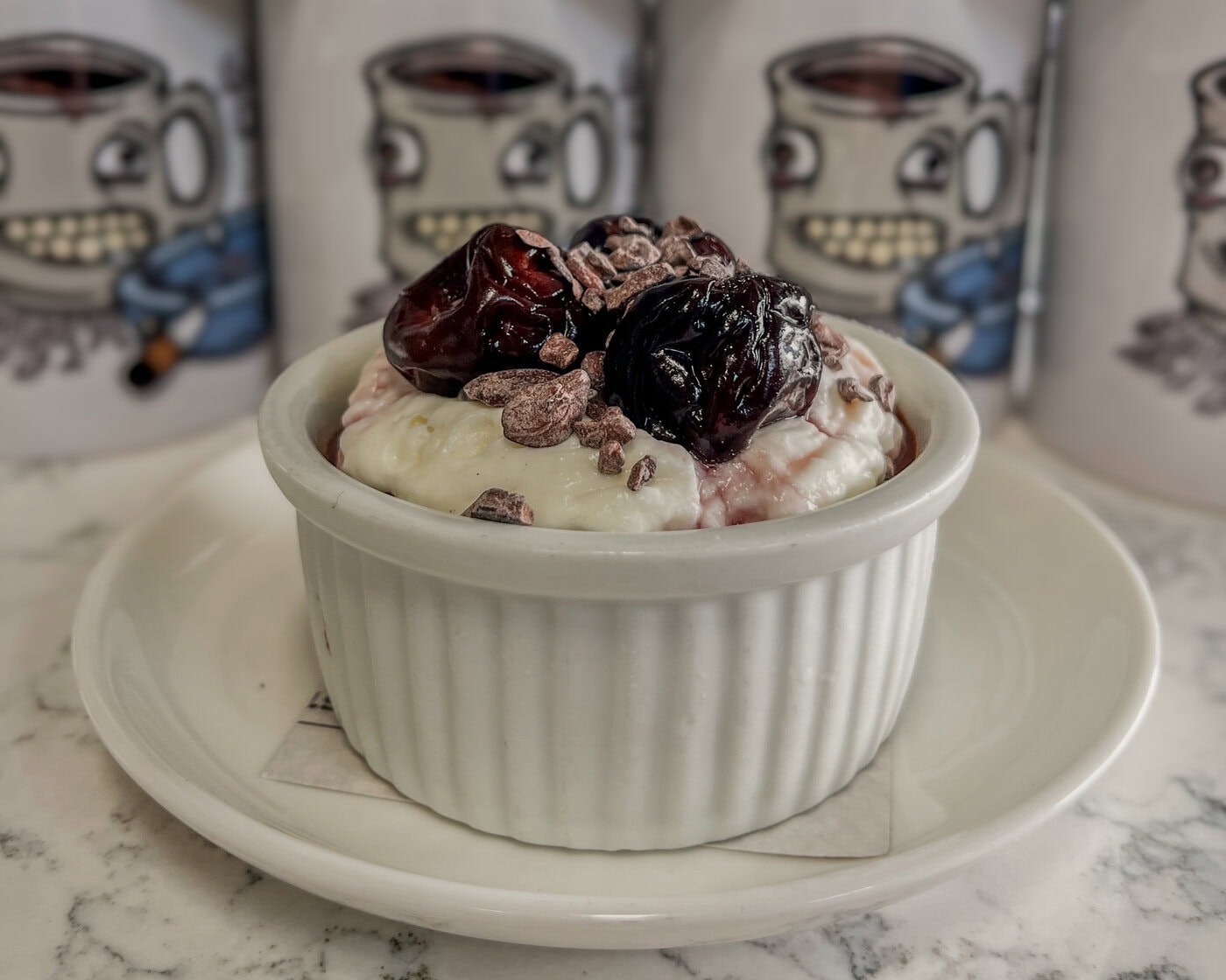 A Small White Ramekin Filled With Creamy Dessert Topped With Cherries And Chocolate Bits Sits On A White Plate, With Illustrated Coffee Mugs In The Blurred Background.