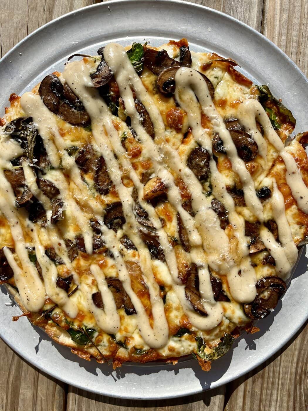 A Baked Pizza Topped With Mushrooms, Spinach, And Melted Cheese, Drizzled With A Creamy Beige Sauce, Served On A Round Metal Plate Atop A Wooden Table In Sunlight.