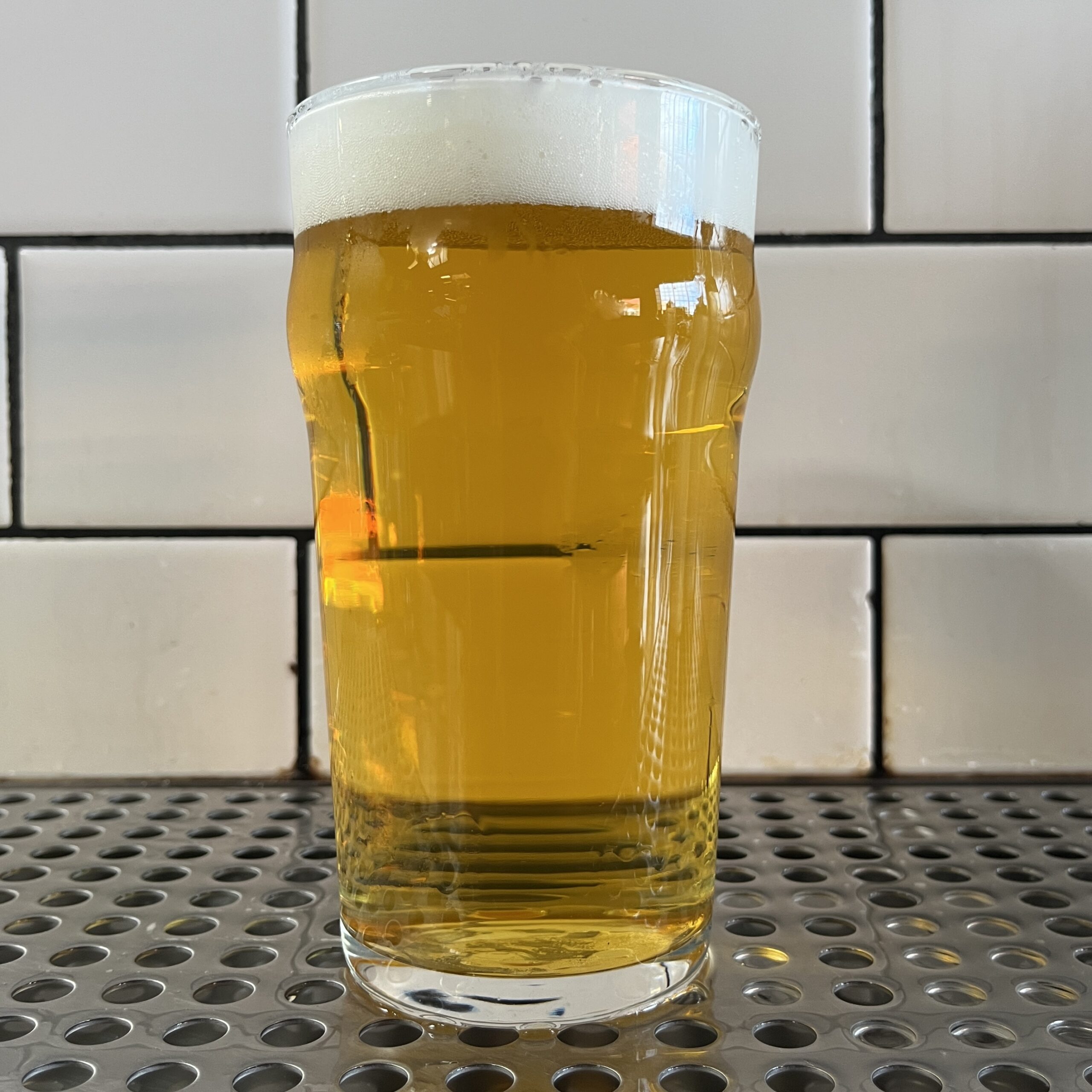A Pint Glass Filled With Golden Beer Topped With A Frothy White Head, Placed On A Metal Grid Surface In Front Of A White Tiled Wall.