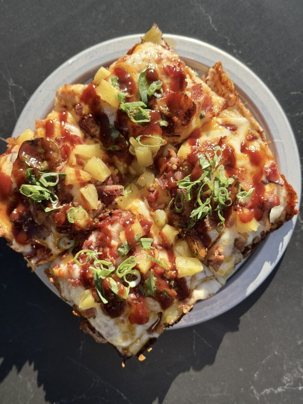 Square-Cut Pizza Topped With Melted Cheese, Pineapple Chunks, Bacon Bits, Barbecue Sauce, And Sliced Green Onions, Served On A White Plate Against A Dark Background.