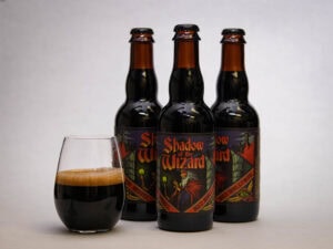 Three bottles of "Shadow of the Wizard" beer with dark fantasy-themed labels stand behind a glass filled with dark beer and a foamy head, all set against a plain light background.