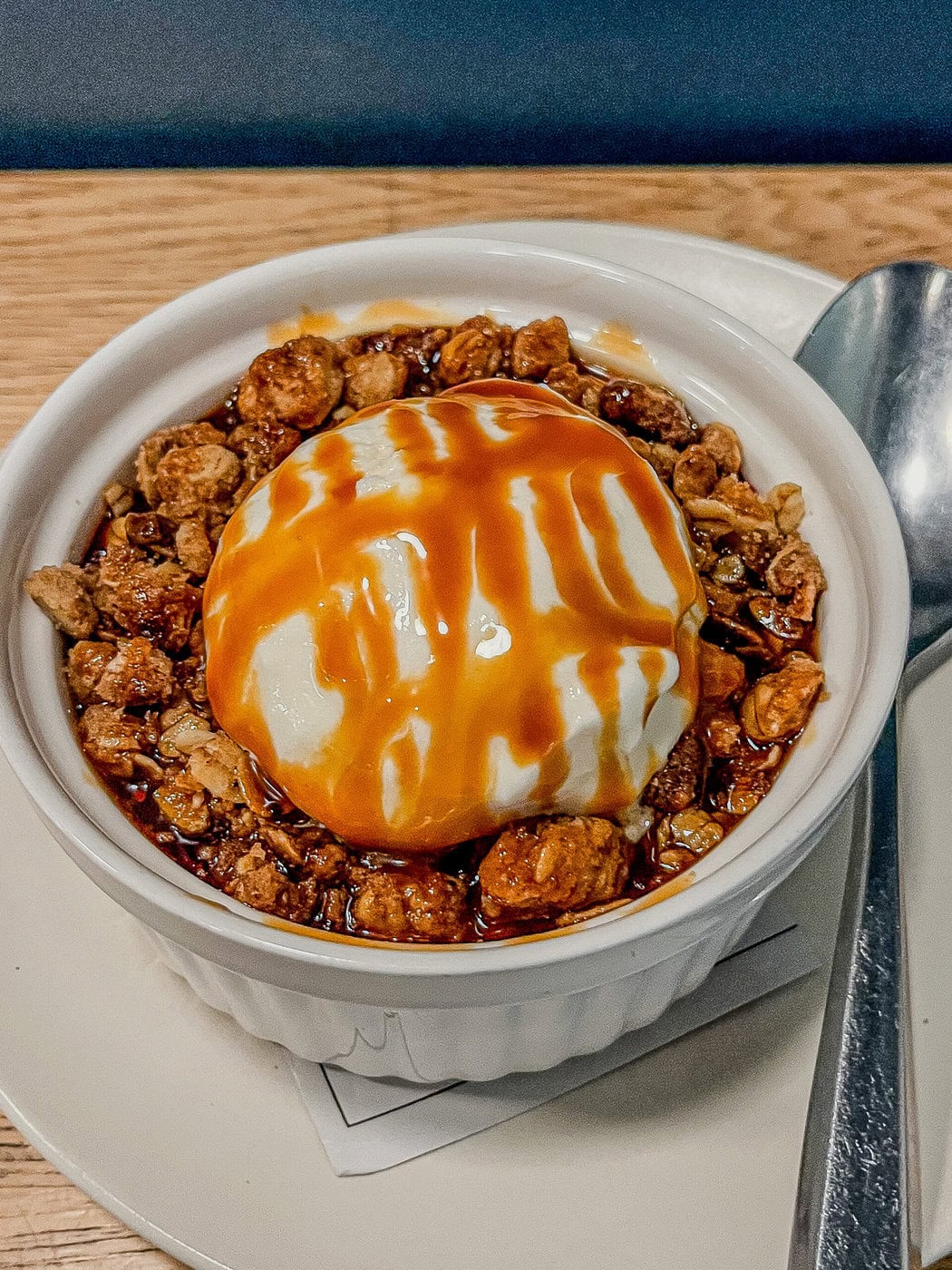 A White Ramekin Filled With Granola, Topped With A Scoop Of Vanilla Ice Cream Drizzled With Caramel Sauce, Sits On A White Plate Next To A Silver Spoon.