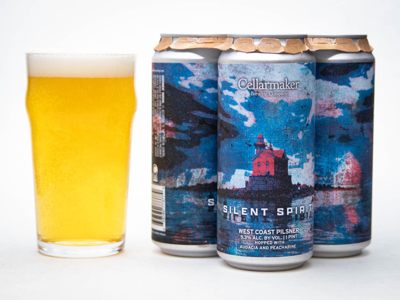 A Full Pint Glass Of Light Golden Beer Sits Next To Three Cans Labeled &Quot;Cellarmaker Silent Spirit&Quot; With Blue And Red Artwork Featuring A Lighthouse And Ocean Scene. The Cans Are 16 Oz West Coast Pilsner.