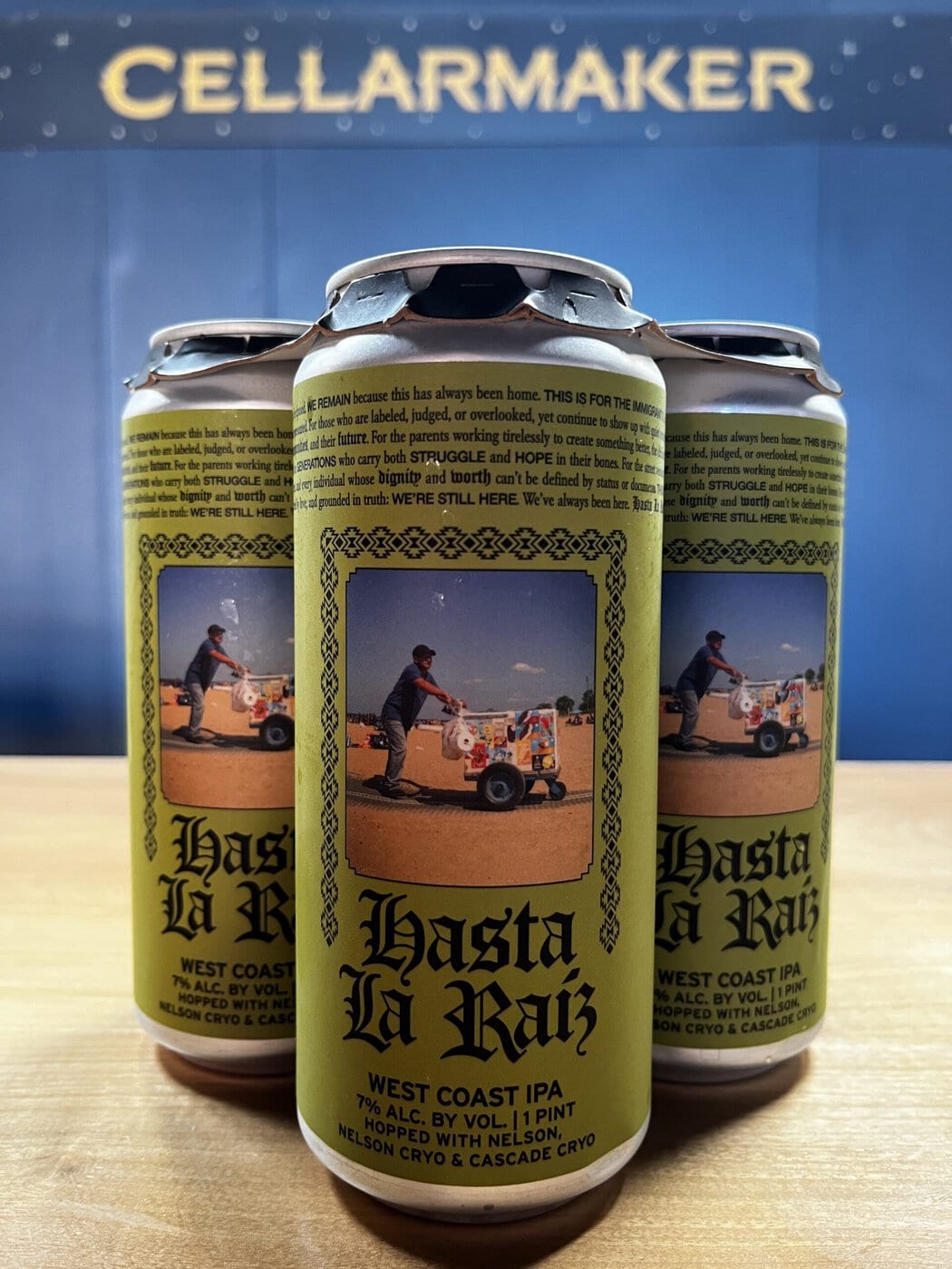 Three Cans Of &Quot;Hasta La Raiz&Quot; West Coast Ipa Sit On A Wooden Surface. The Cans Feature A Yellow Label With An Image Of A Man Pushing A Decorated Cart And The Words &Quot;Cellarmaker&Quot; On A Blue Background Behind Them.