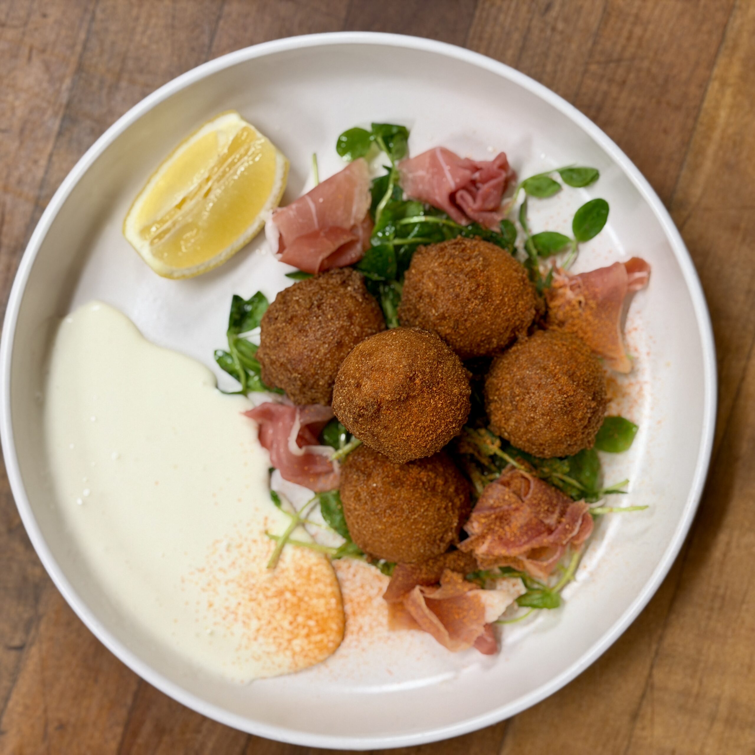 A White Plate With Five Golden-Brown Croquettes On Greens, Slices Of Cured Meat, A Lemon Wedge, And Two Creamy Sauces, Set On A Wooden Surface.