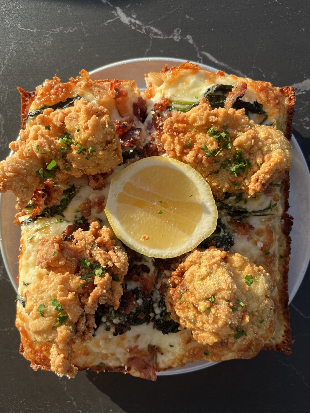 A Square Piece Of Pizza Topped With Four Pieces Of Fried Chicken, Melted Cheese, Greens, Herbs, And A Lemon Wedge, Served On A White Plate On A Dark Surface.
