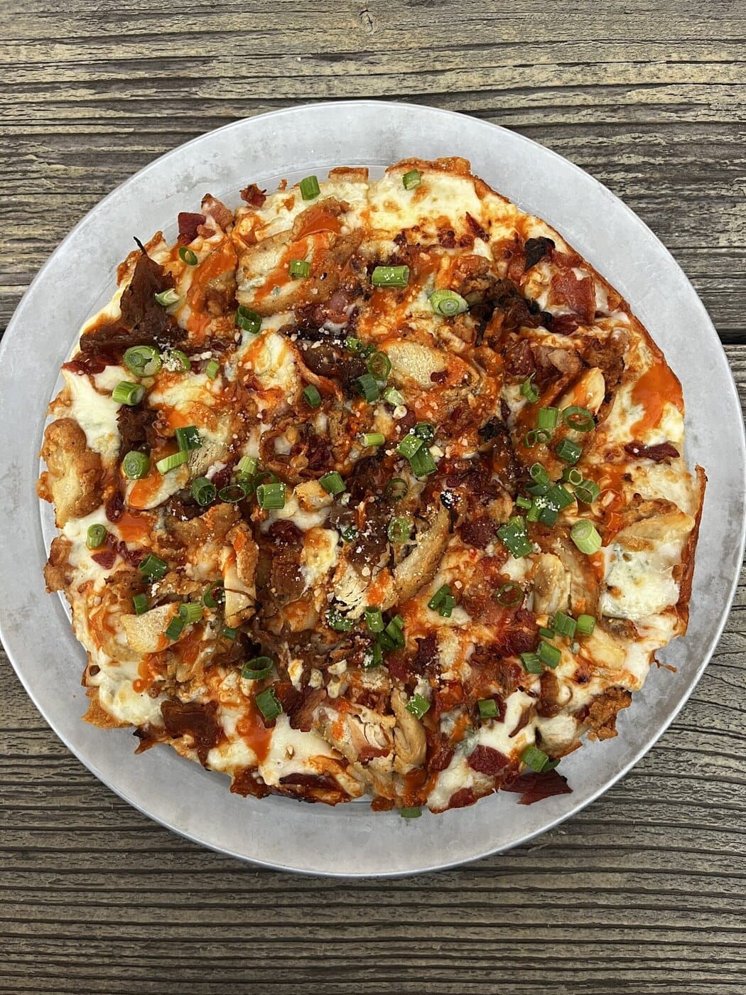 A baked pizza topped with melted cheese, grilled chicken, bacon bits, chopped green onions, and a drizzle of sauce, served on a round metal tray over a rustic wooden table.