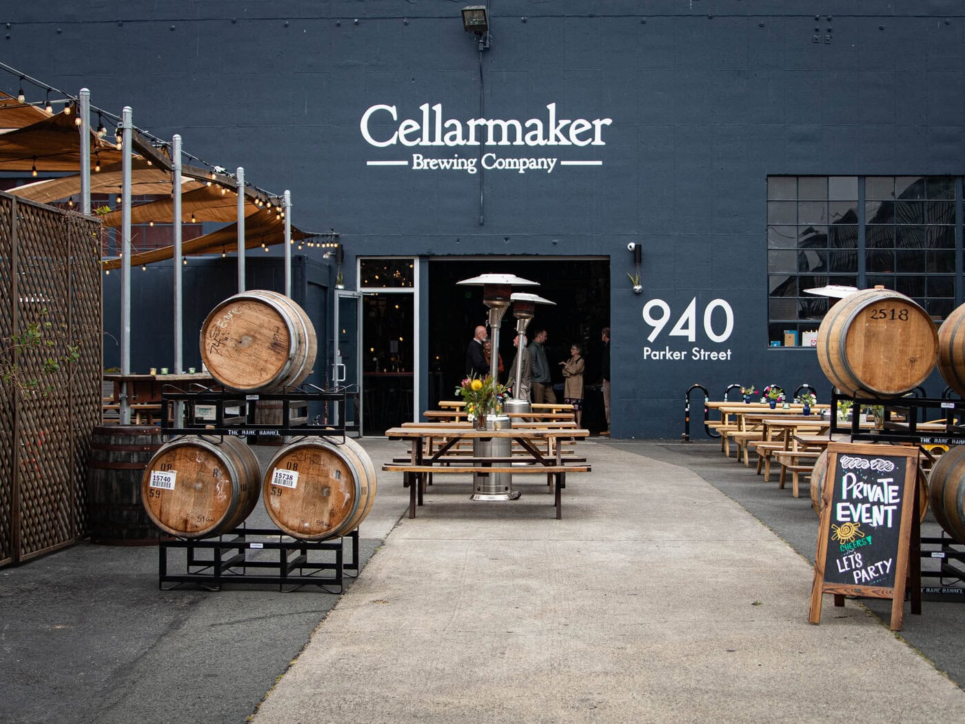 The Entrance To Cellarmaker Brewing Company At 940 Parker Street, Featuring Wooden Barrels, Outdoor Seating With Benches, String Lights, And A Sign Indicating A Private Event.
