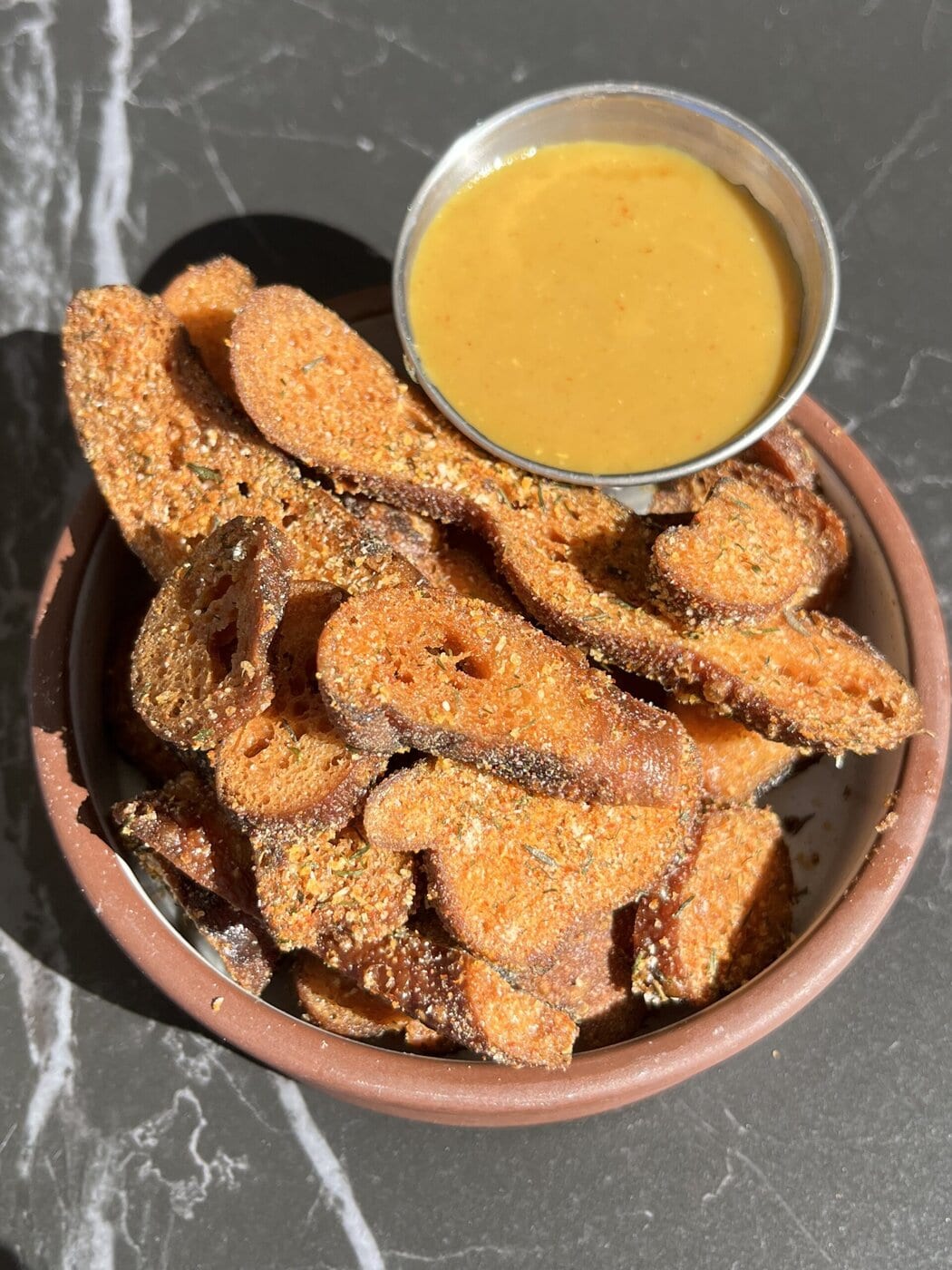 A Bowl Of Crispy, Seasoned Sweet Potato Chips Is Served With A Small Dish Of Golden, Creamy Dipping Sauce On A Dark Countertop.