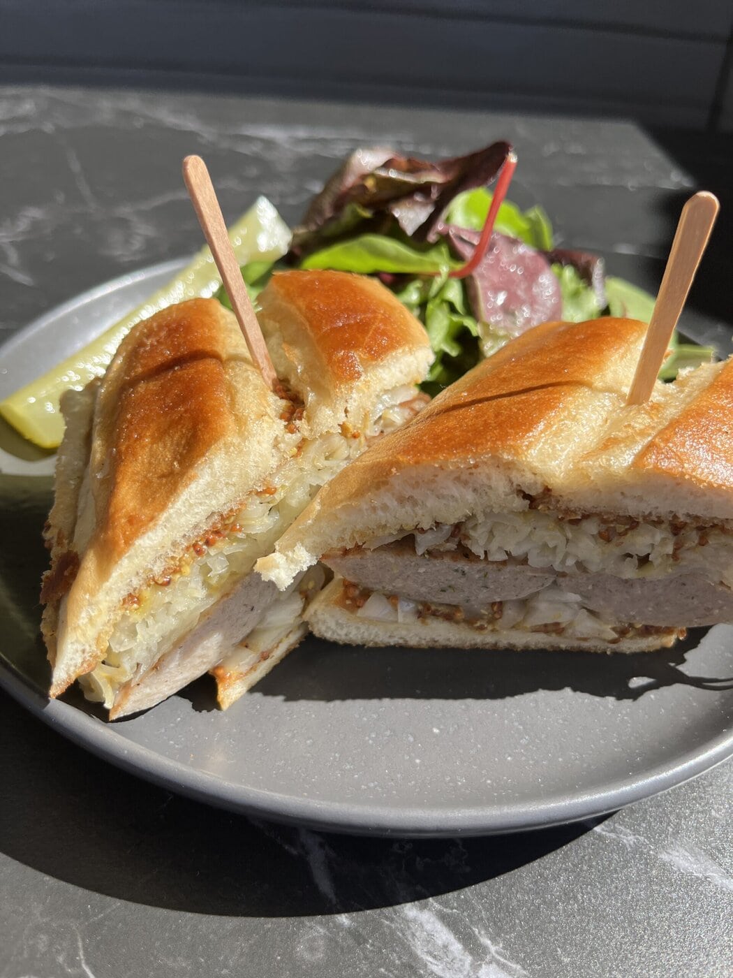 A Close-Up Of A Toasted Sandwich Cut In Half, Filled With Meat And Possibly Sauerkraut, Held Together With Wooden Picks, Served On A Gray Plate With Mixed Greens And A Pickle On The Side.