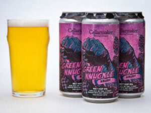 A glass of hazy yellow beer sits next to four cans of Cellarmaker Brewing Company's Green Knuckle Krush, featuring vivid pink and blue artwork with a fist on the cans.