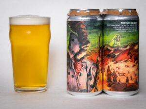 A pint glass of hazy golden beer sits beside two colorful cans with surreal, sci-fi artwork labeled "Foreign Object - WC IPA - TDNE Collab.