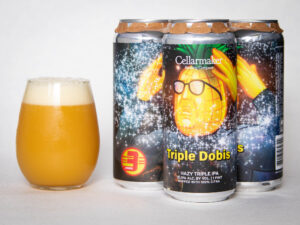 Three cans of Foreign Object - WC IPA - TDNE Collab, with a bright illustrated label, stand next to a glass of hazy, golden beer topped with a frothy head.