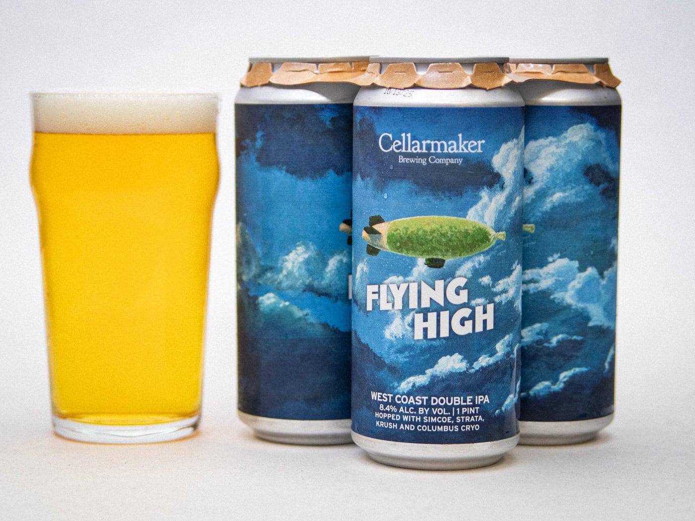 A Pint Glass Filled With Hazy Golden Beer Sits Beside Four Cans Of Cellarmaker Brewing Company'S &Amp;Quot;Flying High&Amp;Quot; West Coast Double Ipa, Which Features A Zeppelin Flying Among Clouds On The Blue Label.