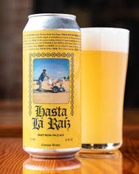 A Can Of Hasta La Raiz Hazy Ipa, Featuring A Label With A Man Pushing A Cart, Stands Next To A Glass Filled With Hazy Pale Beer On A Wooden Surface.