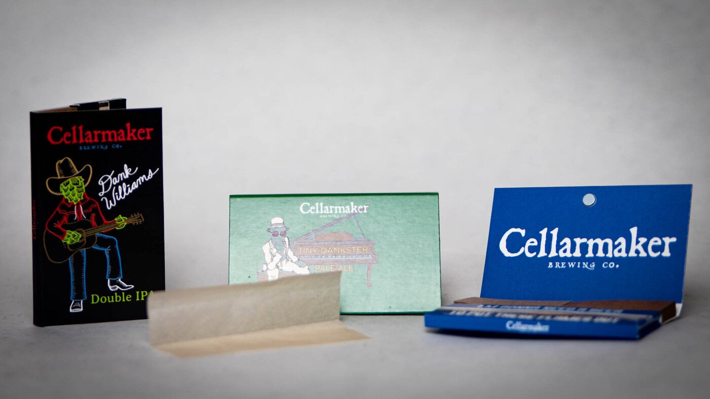 Three Packs Of Rolling Papers Are Shown On A White Surface, One Open With A Sheet Partially Pulled Out. The Packaging Features Text And Illustrations, Including A Figure Playing Guitar.