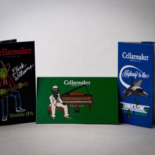 Three colorful Rolling Papers beer cans are shown: one with a green skeleton cowboy, one with a skeleton playing piano, and another with a fighter jet. Each features playful designs and bold typography.