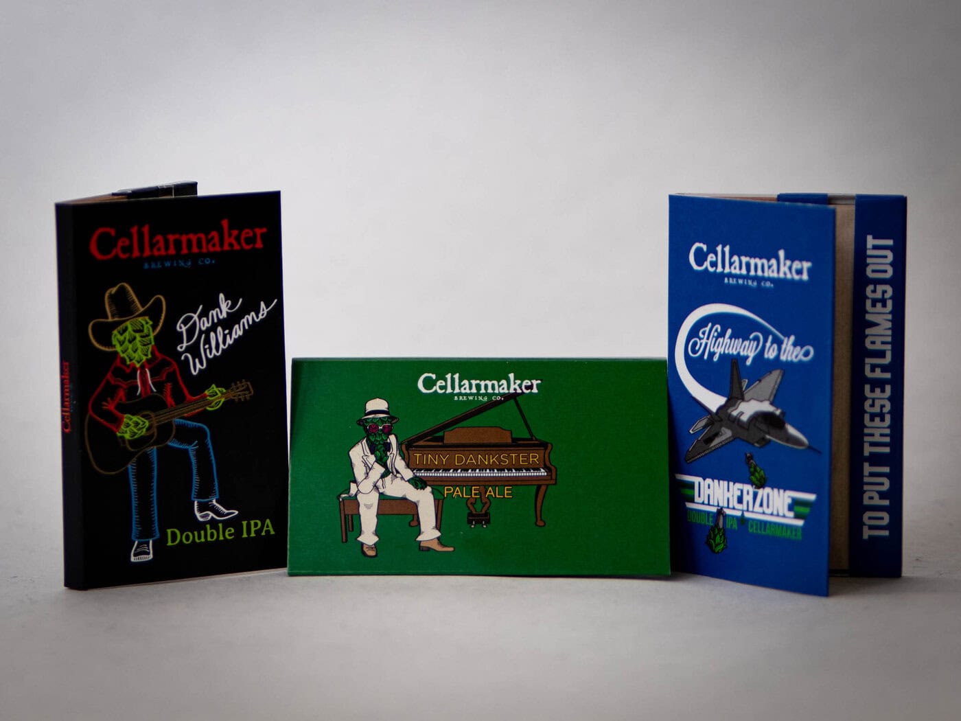 Three colorful Rolling Papers beer cans are shown: one with a green skeleton cowboy, one with a skeleton playing piano, and another with a fighter jet. Each features playful designs and bold typography.